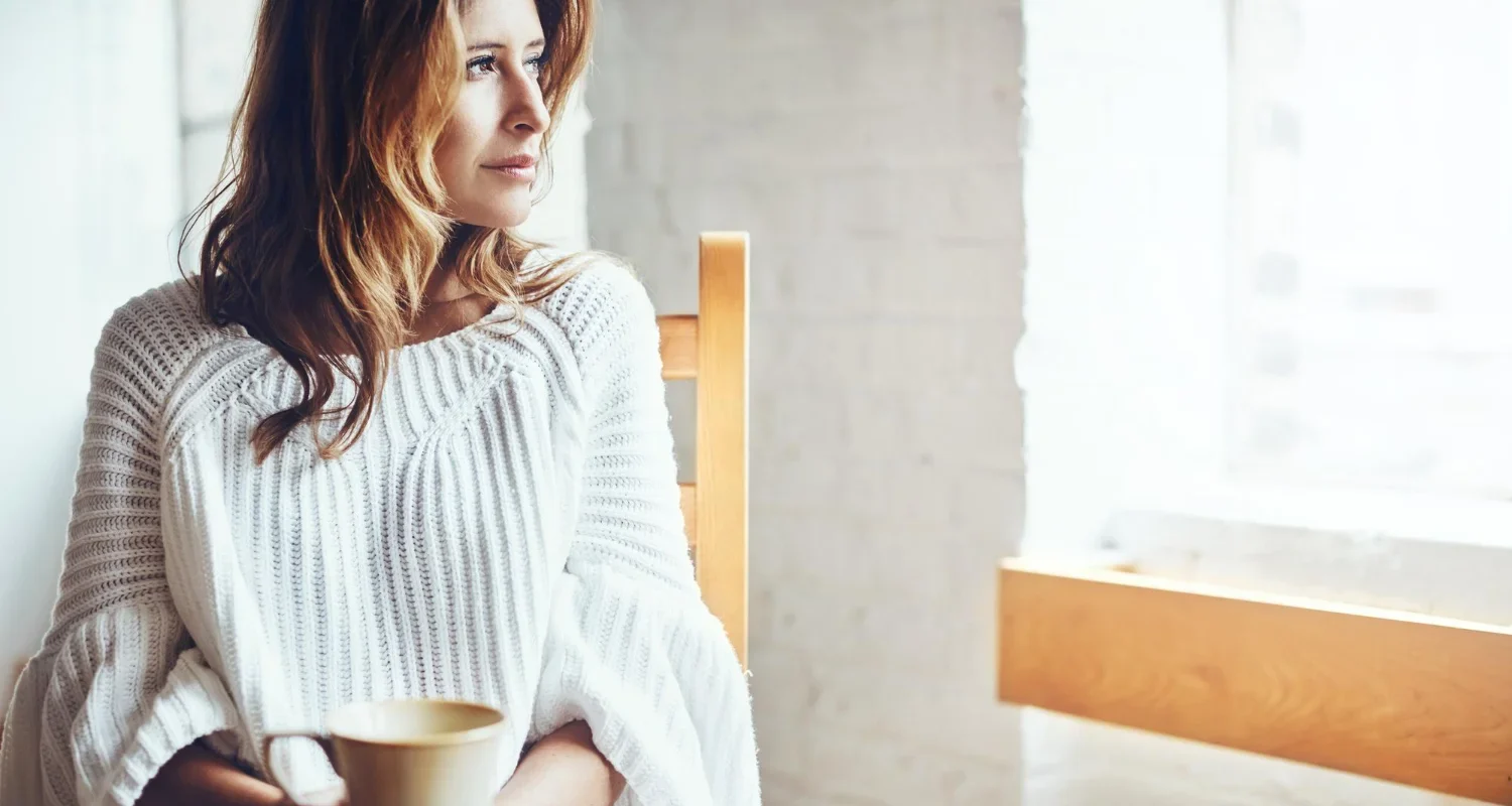 Woman in her 40s reflecting on her health and fitness journey while looking out a window at home