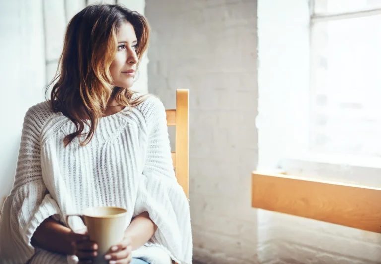 Woman in her 40s reflecting on her health and fitness journey while looking out a window at home