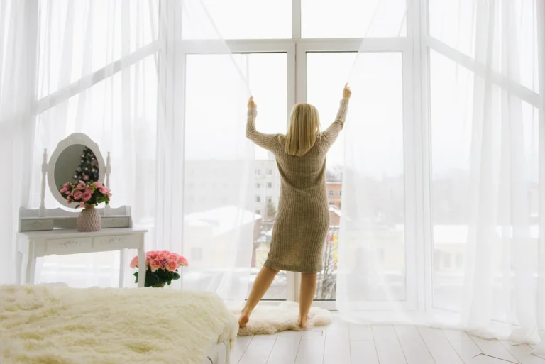 Woman in cream activewear stretching arms overhead in a bright sunlit home workout space