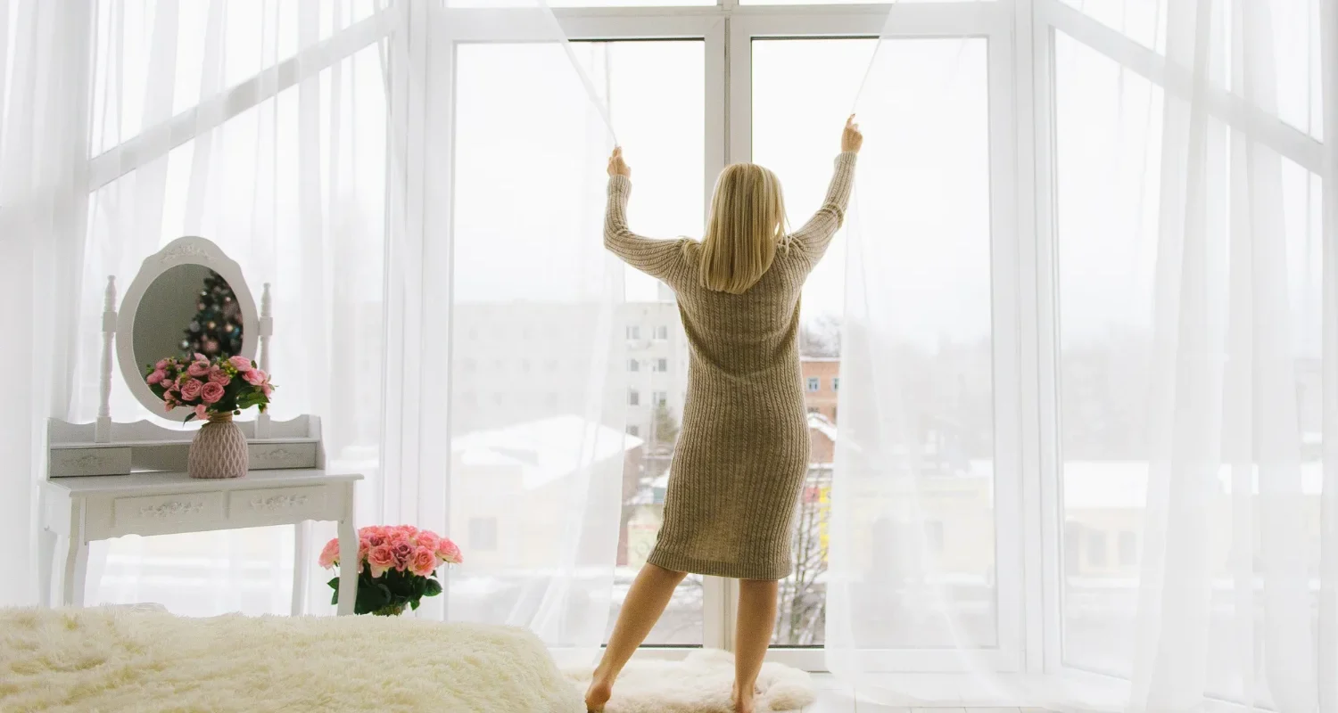 Woman in cream activewear stretching arms overhead in a bright sunlit home workout space